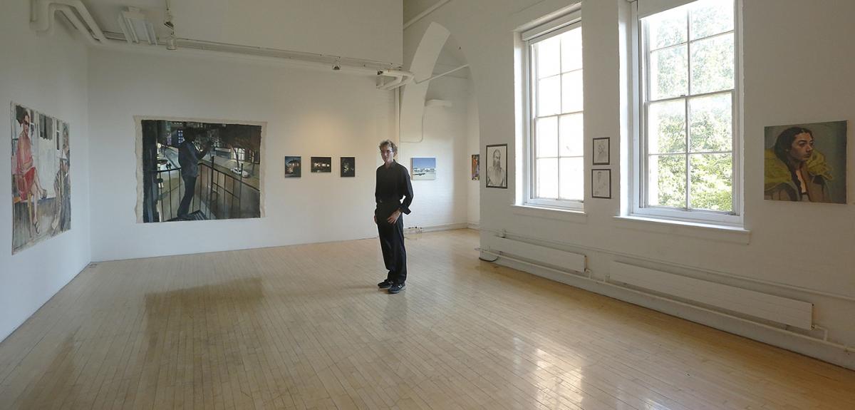 young man dressed in black standing in a gallery with large windows and wooden floors