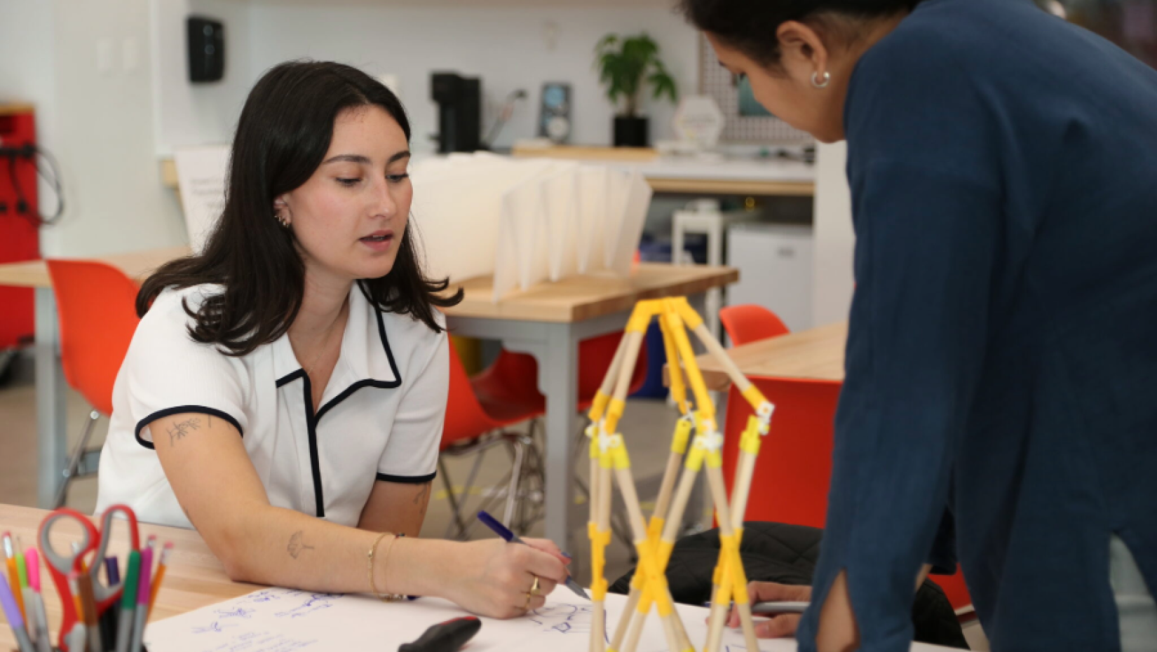 Two people collaborate in a design studio, sketching ideas beside a small structural model.