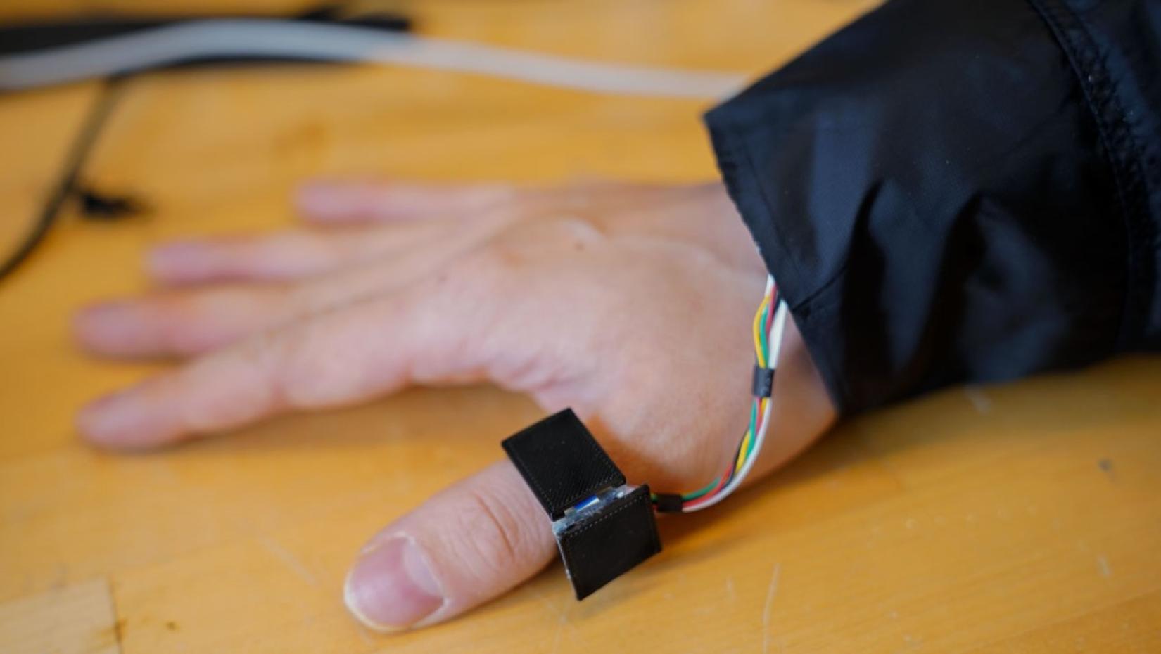 A hand pressed to a table. Around the thumb is an electronic device.