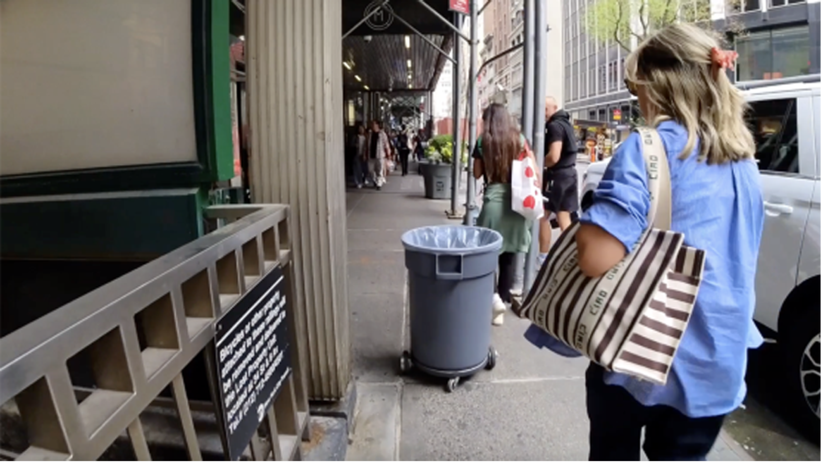 A woman walks down a street following a trash can on wheels.