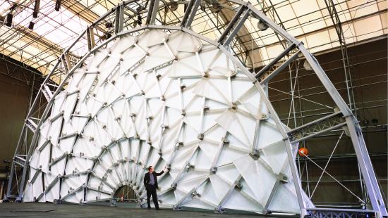 A person stands in front of a large structure in the shape of a semi-circle made by multiple articulated geometric pieces.