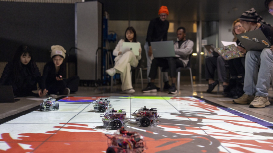 Design Tech students sit around a floor mat that is occupied by five robotic vehicles.