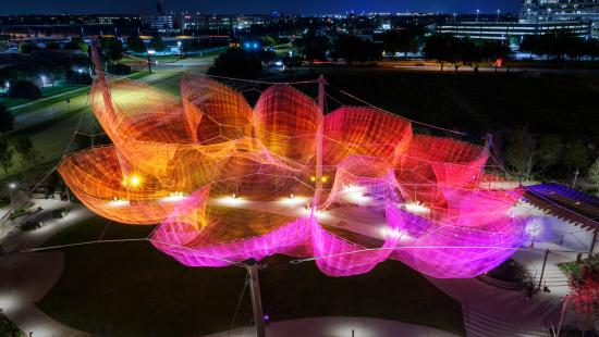 A big sculpture that resembles an open flower made of fabric hanging above an open park and illuminated with coloful lights in the night.