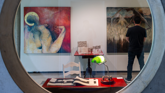 Interior view of Bibliowicz Family Gallery as seen from the porthole window, with two large acrylic paintings in the center and a red desk with an open book and green lamp in the foreground.