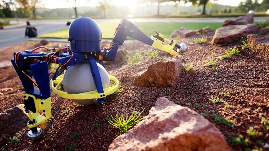 A mechanism with robotic arms made of metal and plastic parts crawling among a rocky field.